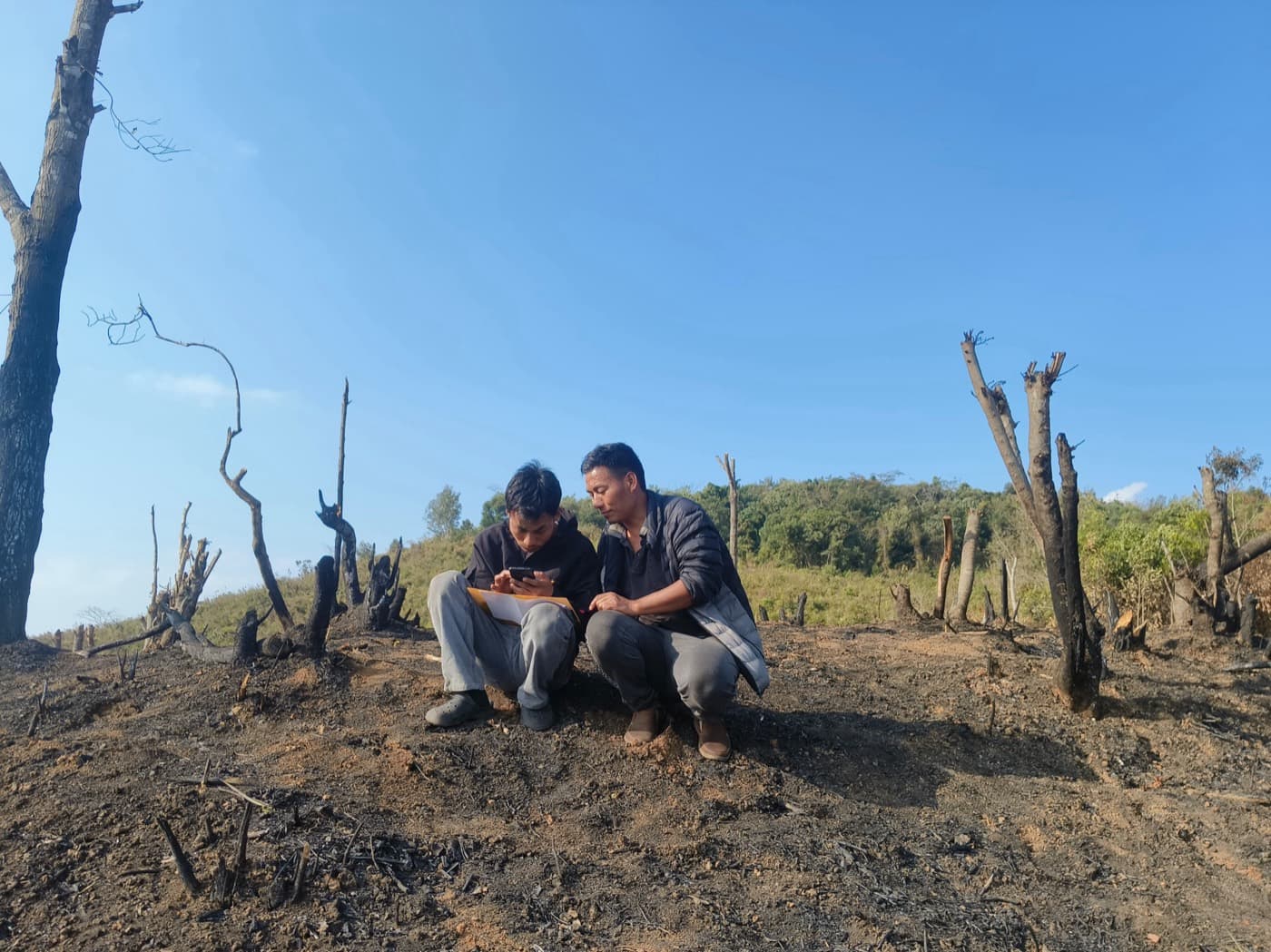 Two field surveyors crouched on a cleared hill slope in Nagaland, recording plot measurements on a phone and a paper form.